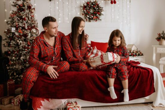 Father, mother and daughter all in matching holiday pajamas. The young daughter is opening a present while the parents look on.